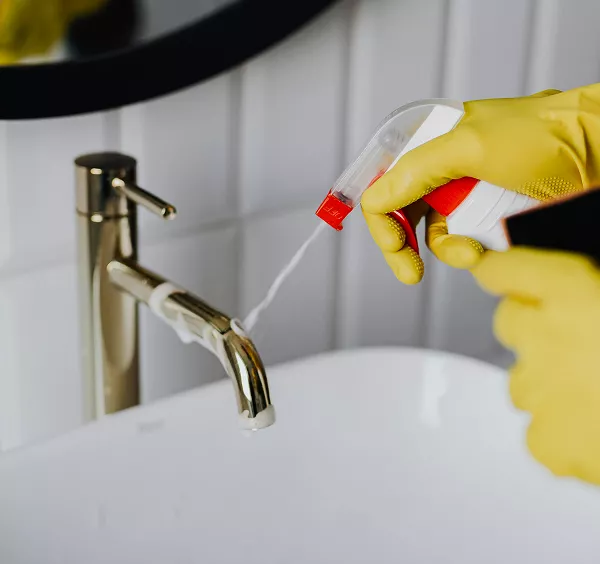 Close-up of a person wearing yellow gloves spraying cleaner onto a gold-colored bathroom faucet and white sink basin.