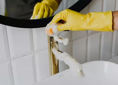 A close-up of a hand in a yellow rubber glove scrubbing a golden bathroom faucet with a foamy sponge near a white sink and tiled wall.