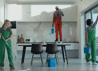 Three professional cleaners in green and red overalls working in a modern kitchen, mopping the floor, and cleaning high walls and windows.