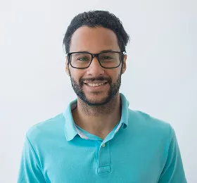 Smiling Black man with glasses wearing light blue polo shirt, photographed against white background in professional headshot style portrait