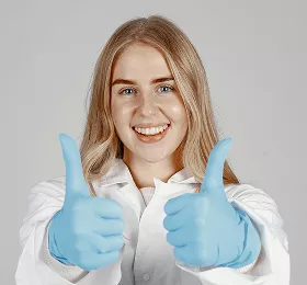 Young blonde woman with bright smile wearing white shirt and blue gloves, giving enthusiastic double thumbs up gesture against gray background