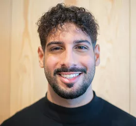 Young man with dark curly hair and beard wearing black sweater, smiling warmly at camera in casual indoor portrait with wooden background
