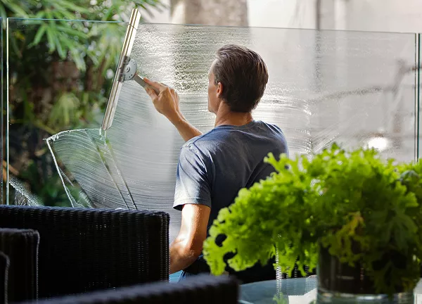 A man from behind is using a squeegee to clean a large glass pane or barrier, likely outdoors or on a patio, with a potted plant in the foreground.