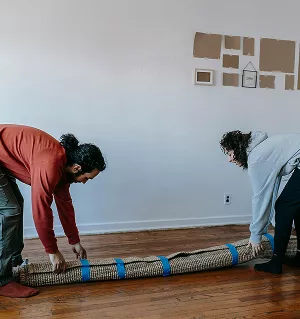 Two people are rolling up a large, woven area rug on a wooden floor, securing it with blue straps. This suggests a cleaning preparation or moving task.